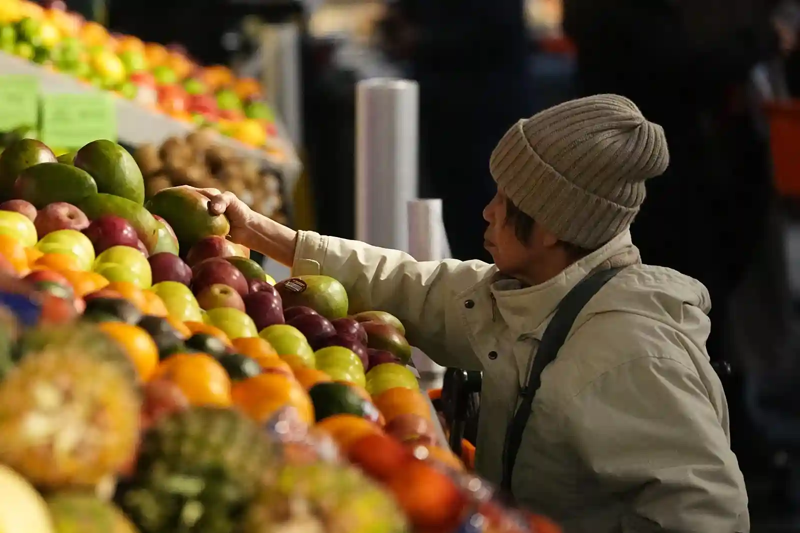 An older shopper wearing a winter hat and coat reaches for apples at a grocery store produce display filled with colorful fruits.