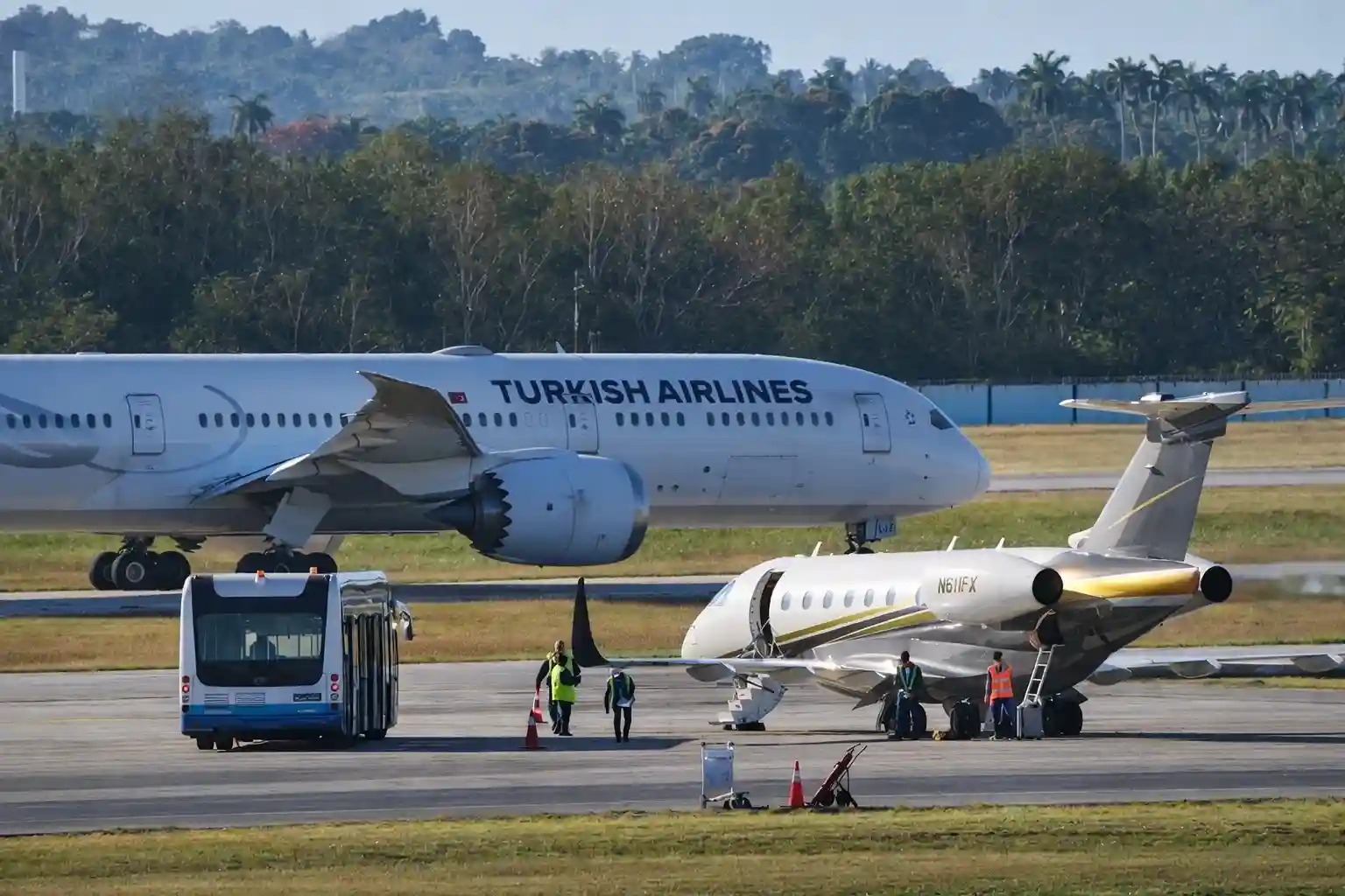 Turkish Airlines jet taxiing on the airport runway while a private jet is serviced nearby.