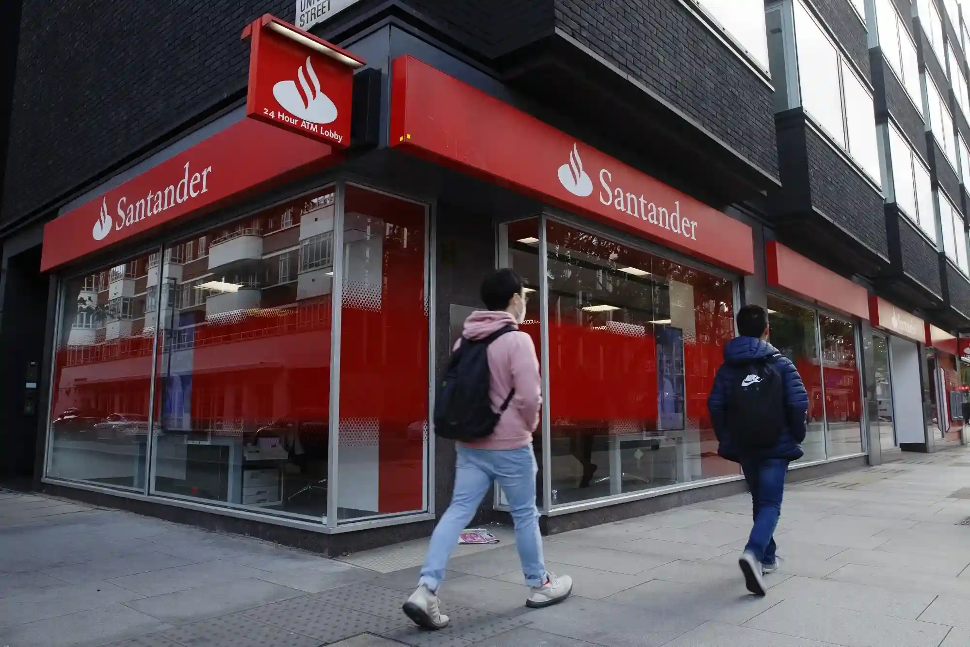 Santander bank storefront with '24 Hour ATM Lobby' sign, people walking past the bank on the sidewalk.