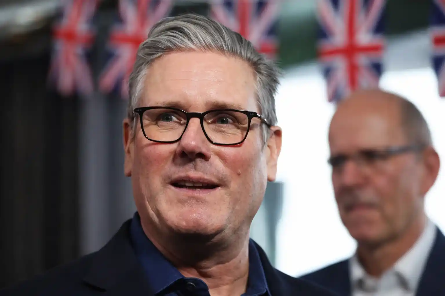 Keir Starmer, leader of the Labour Party, speaks to the press amidst political tensions, wearing a suit in front of British flags.