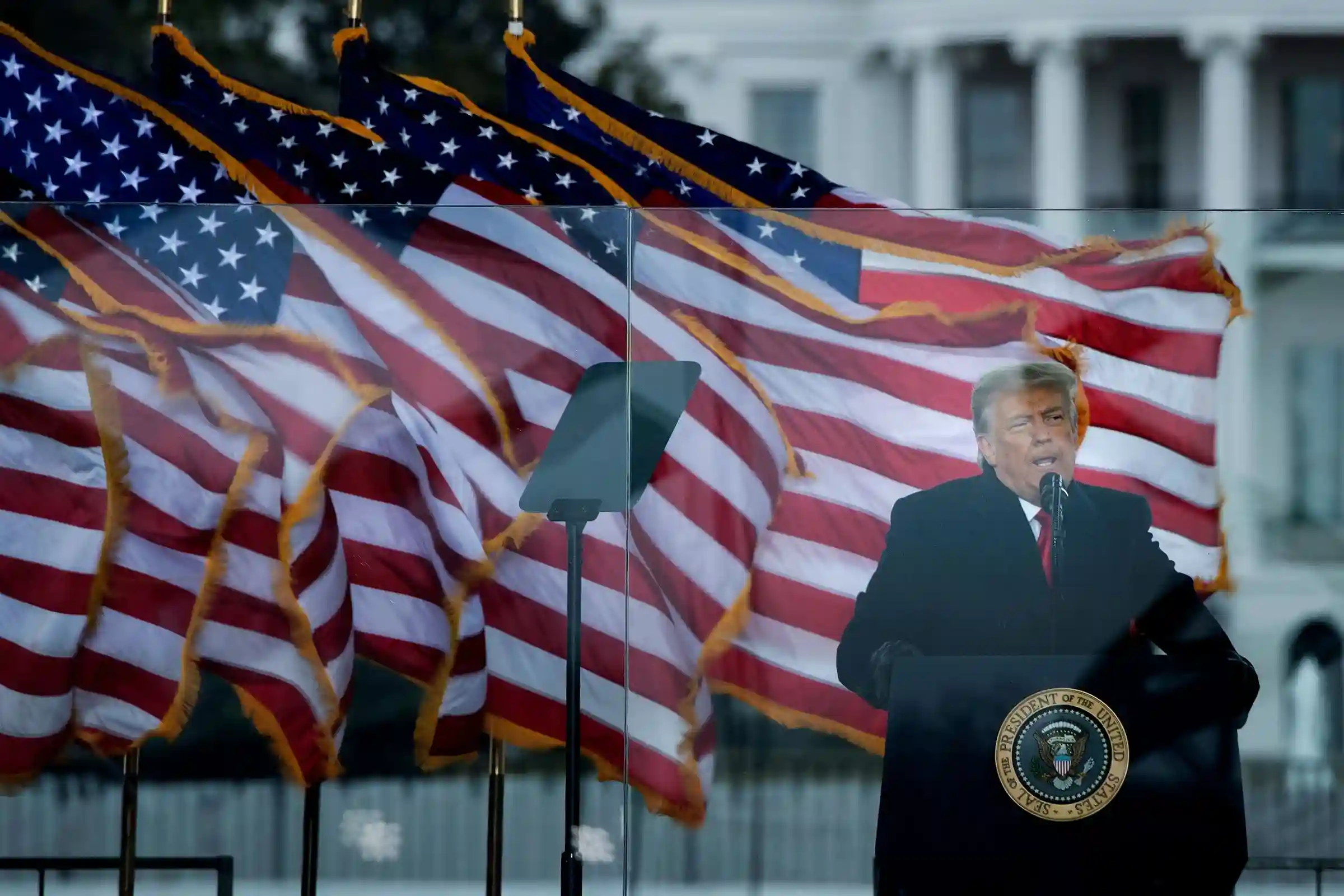 Donald Trump delivering a speech with multiple US flags in the background at a public event.