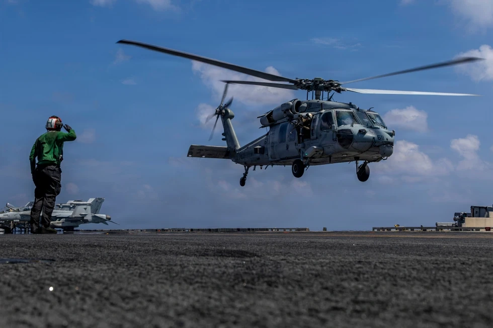 A US Navy helicopter taking off from the deck of an aircraft carrier, with a crew member signaling from the deck.